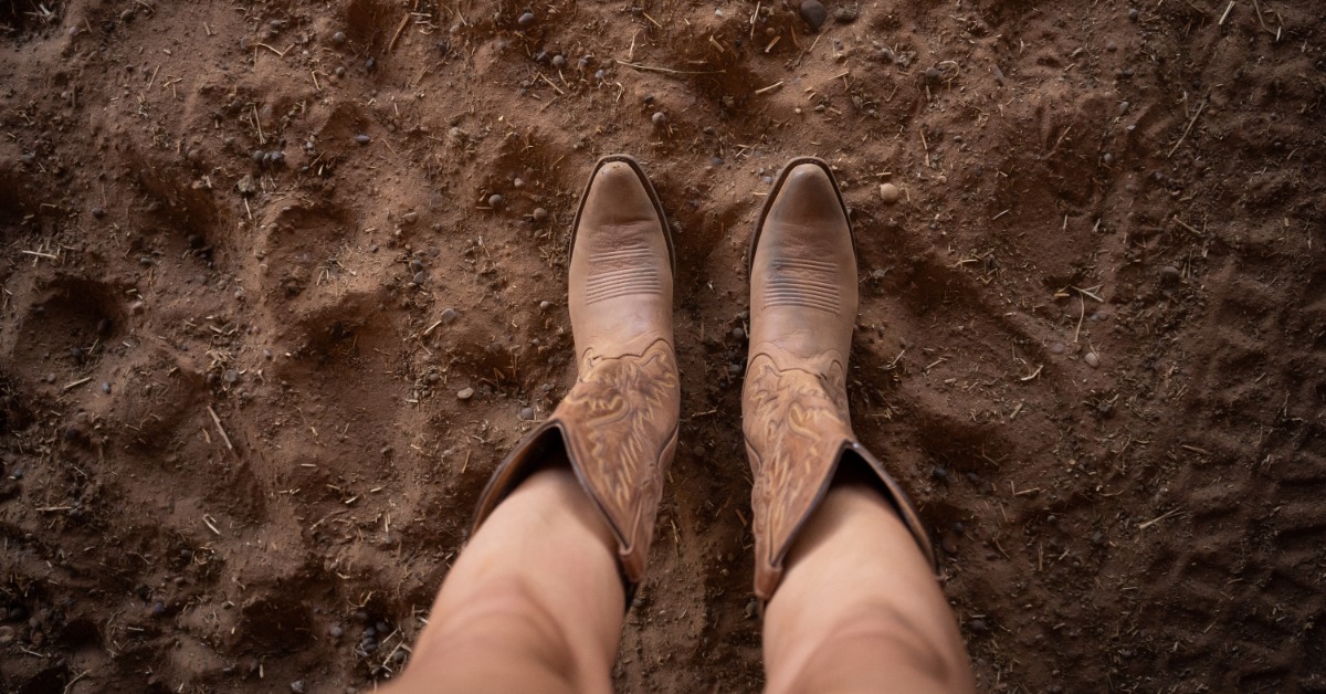  pair of tan cowboy boots worn by a person standing on reddish dirt with scattered stones and straw.