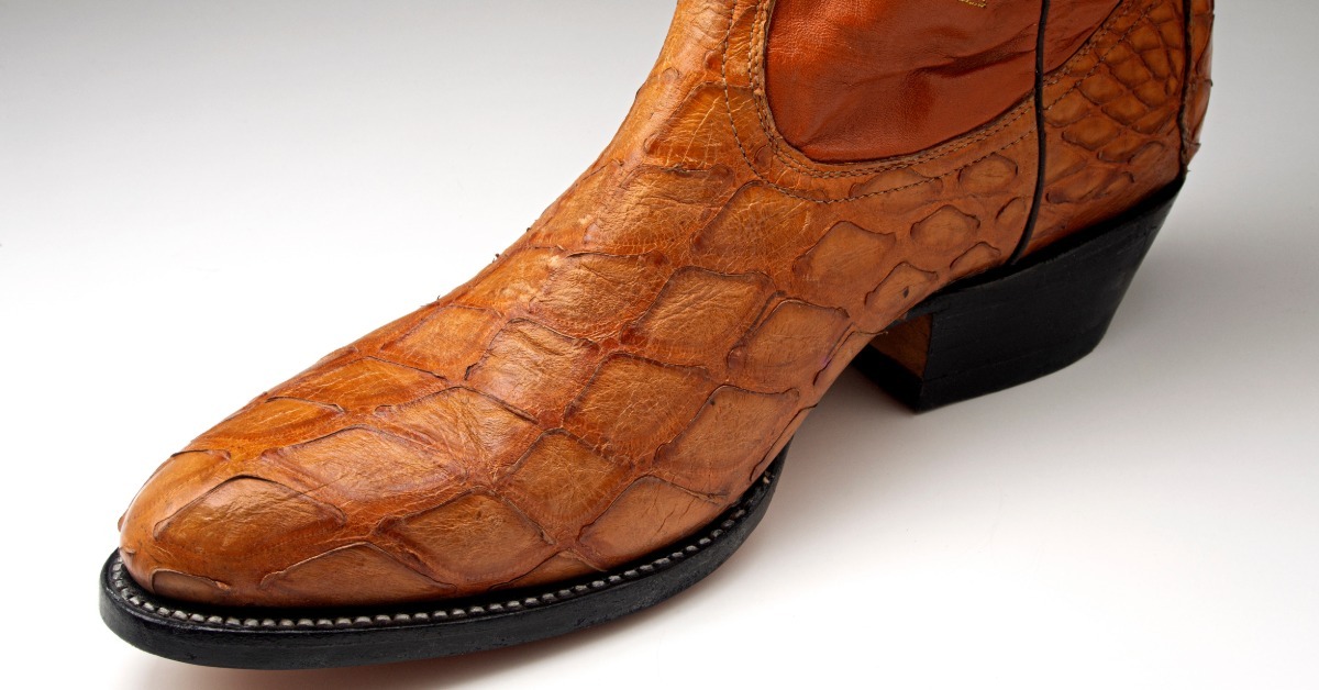 A brown cowboy boot resting on a light surface, showing raised scale texture, stitching, and a black heel.