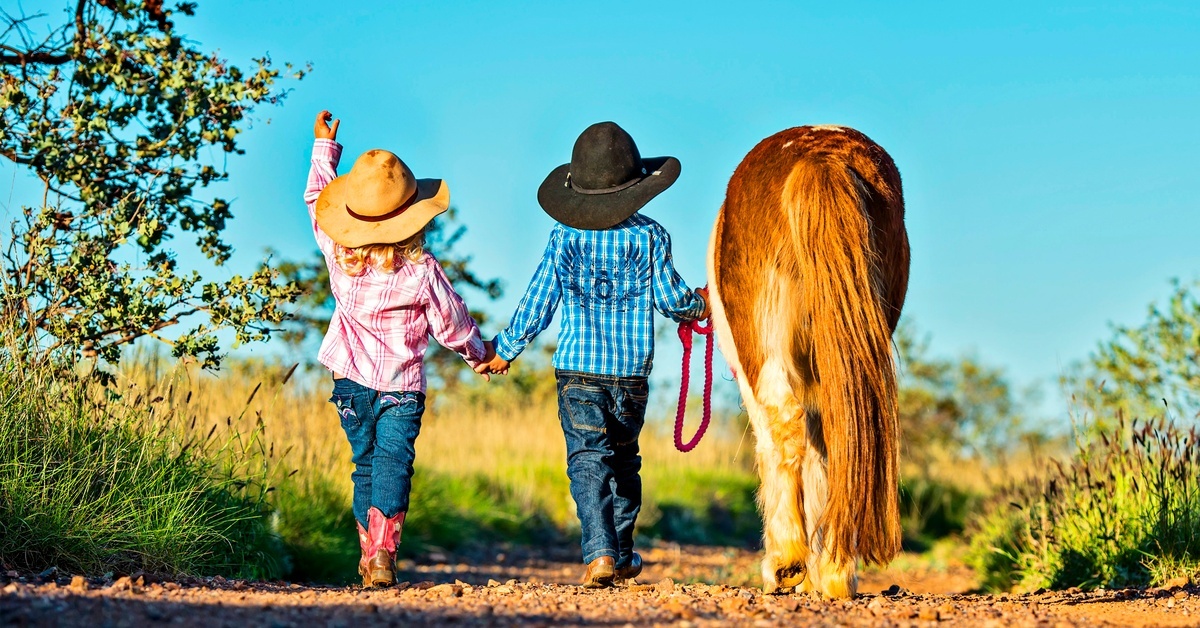 Two children in cowboy hats walking down a dirt path beside a pony, with grass, trees, and blue sky nearby.