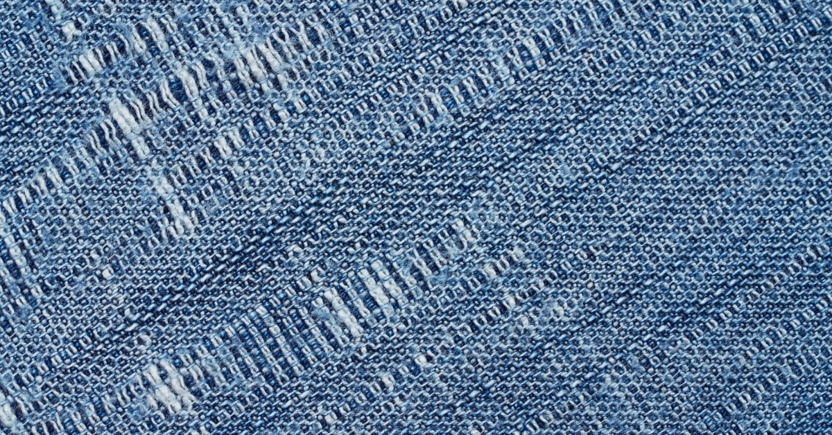 Blue woven denim fabric with diagonal threads, light worn patches, and varied blue and white texture.