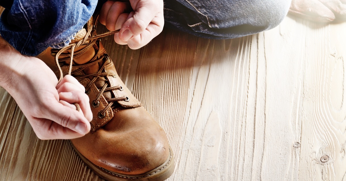 A man in blue jeans kneeling on a wooden floor and tying the laces of a light-brown work boot.