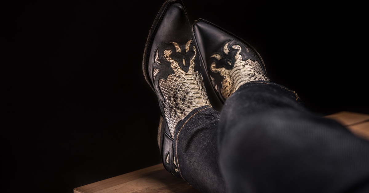 Someone in black jeans resting their feet up on a wooden surface. They are wearing black boots with a tan snakeskin design.
