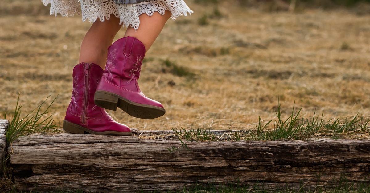 Everything You Need To Know About Kids’ Cowboy Boots A young girl wearing reddish-brown cowboy boots running on a log while in the middle of a field with dead grass.